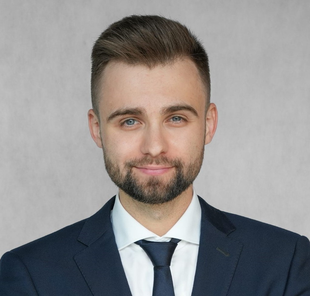 Mężczyzna w ciemnym garniturze i krawacie w paski. Ma krótkie, ciemne włosy i odrobinę zarostu. Patrzy wprost, na neutralnym tle.