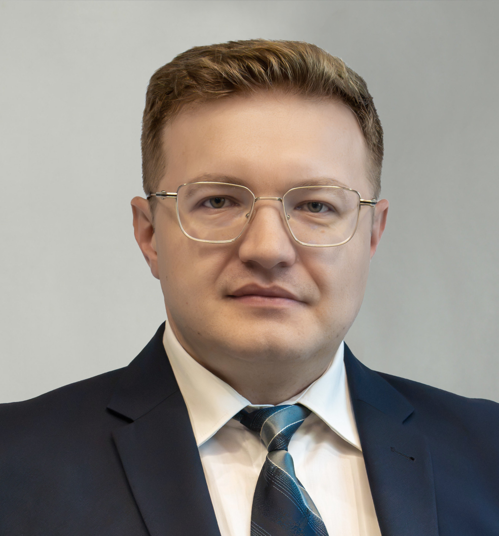 Mężczyzna w ciemnym garniturze i krawacie w paski. Ma krótkie, ciemne włosy i odrobinę zarostu. Patrzy wprost, na neutralnym tle.