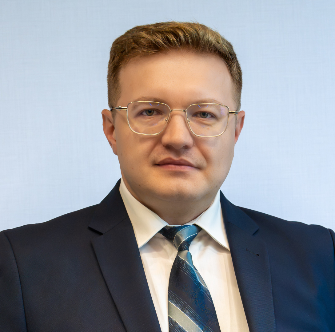 Mężczyzna w garniturze z krawatem w paski, o krótkich ciemnych włosach i brodzie, na neutralnym tle.